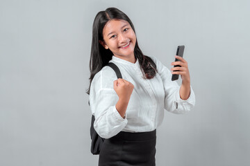 Fototapeta premium Happy Asian teenage student in school uniform showing success gesture while holding smartphone. Concept of back to school, mobile learning, achievement, and digital education.