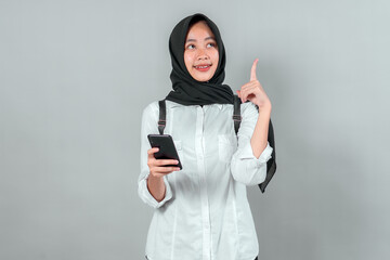 Muslim teenage girl in hijab and white shirt holding smartphone while looking up and pointing with...
