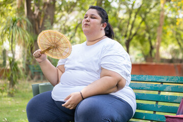 Plus size indian woman using hand fan in hot summer day. Overweight lady suffering from heat wave...