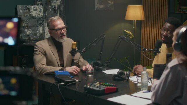Handheld shot of mature Caucasian entrepreneur or writer in formalwear giving interview to diverse hosts while camera recording in podcast studio