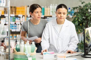 Professional asian female parmacist working computer and helping young girl client with prescription in hand look for medicine in modern drugstore