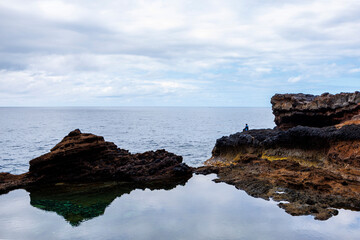 Solitary figure seated on rugged volcanic coastline gazing at the sea under a cloudy sky, evoking...