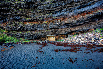 Small wooden structure nestled against dramatic layered volcanic cliffs and surrounded by black sand, illustrating scale, solitude and natural resilience.