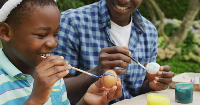 Animation of happy african american father and son painting easter eggs together in garden