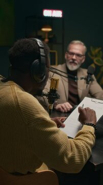Vertical shot of young black man reading script and asking mature Caucasian guest questions during podcast recording in studio