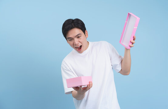 Young man excitedly opening a pink gift box against a light blue background in a cheerful indoor setting - Powered by Adobe