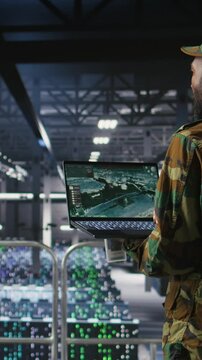 Vertical video Military technician reviewing tactical dashboard on laptop screen inside secure server hub. Army expert using strategic communications and infrastructure oversight software on notebook