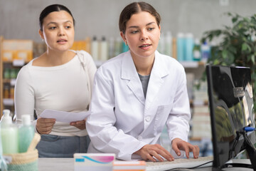 Young professional female parmacist working computer and helping asian female client with prescription in hand look for
