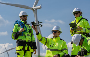 Fototapeta premium Maintenance engineer team standing at windmills at wind turbine farm. Group of people wear safety helmet and uniform working at alternative renewable energy wind station. Sustainable energy technology