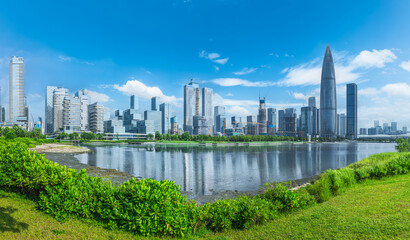 Obraz premium Panoramic vista of Shenzhen's modern cityscape with building reflections in the water, viewed from a lush waterfront park in China.