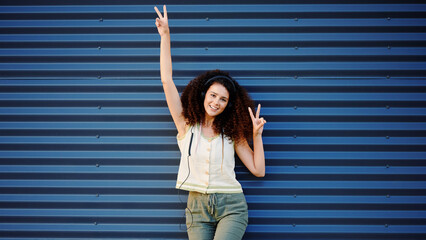 joyful woman with headphones posing against blue corrugated metal wall, making peace signs....