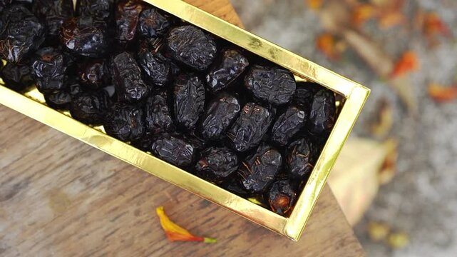 Dried black fruit dates in golden rectangular box on wooden surface with blurred background and small orange flower petal nearby