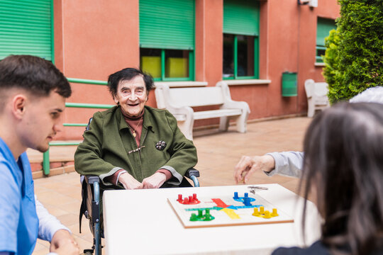Elderly woman enjoying a game of parcheesi with a nurse and friends, promoting social interaction and cognitive stimulation in a senior care facility