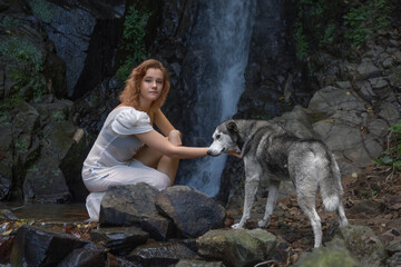 A girl in a transparent long white tunic, sitting against the backdrop of a waterfall in a grotto, scratches her dog's neck