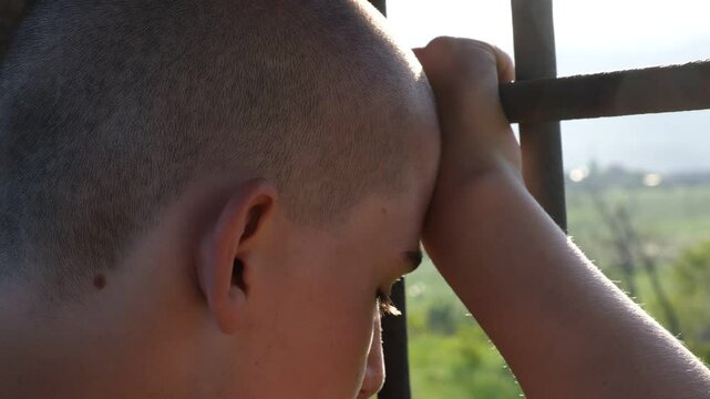 A teenage boy wrongly convicted presses his forehead to the prison bars, gripping them tightly as he looks out with sadness. Juvenile detention, injustice, and lost childhood behind bars.