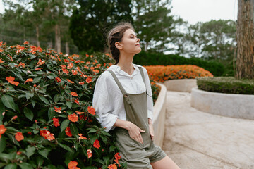 Young woman in casual outfit enjoying a serene moment in a blooming garden with vibrant flowers,...