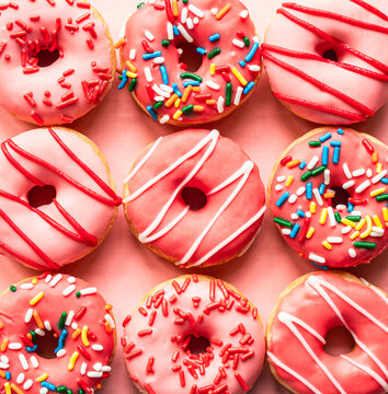 Close up of  9 donuts with pink glaze and sprinkles on pink backdrop.