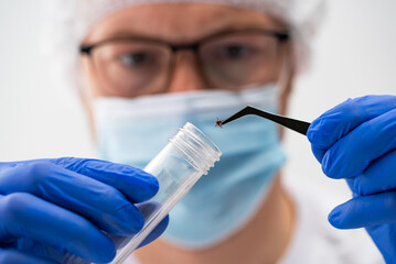 Scientist wearing blue gloves, a white cap, and a face mask uses tweezers to carefully place a tick...