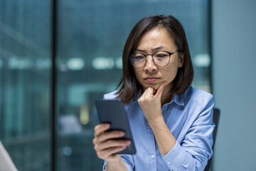 A thoughtful Asian woman wearing glasses examines her smartphone, pondering a message in a modern...