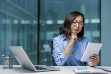 An asian businesswoman is thoughtfully reviewing information on a tablet while sitting at her desk in a modern office setting. There is a laptop.