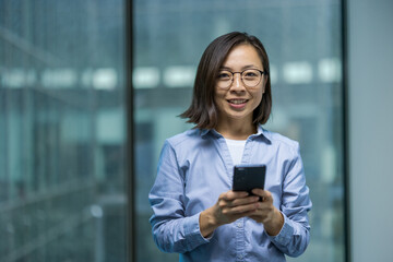 A smiling asian woman holds a smartphone indoors, looking at the camera. She wears glasses and a...