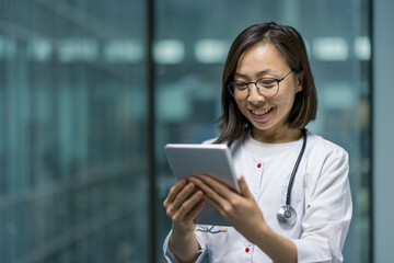 A smiling asian female doctor is using a tablet. The woman is wearing glasses and a stethoscope around her neck.