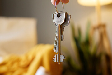 A close-up shows a hand holding a set of house keys on a keyring, with a blurred interior...