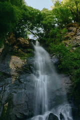 Obraz premium Majestic waterfall cascading down rocky cliffs surrounded by lush green forest, creating a serene and refreshing natural landscape in bright daylight.