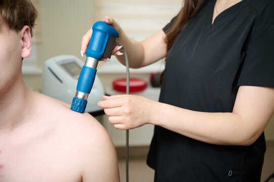 Physical therapy of shoulder with acoustic shock waves. Close up view of doctor performing shock wave therapy procedure on back. Female holding pneumatic shock wave therapy machine for pain relief.