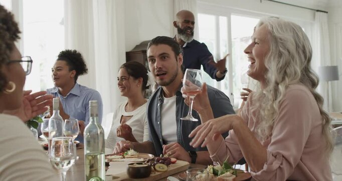 Diverse friends and family sharing dinner at home with wine glasses welcoming newcomer with hug
