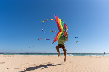 Happy young boy jumping on beach while flying a colorful rainbow kite, enjoying a sunny windy day by the ocean with clear skies and turquoise waves