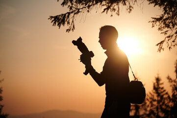 Silhouette of a photographer in the mountains taking pictures at dawn. Tourist holding camera on the mountain hill on sunrise background. Man takes photo of mountain in the rays of the setting sun.