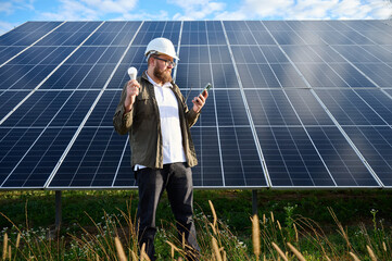 Engineer in uniform with protective helmet near solar station holding light bulb. Supervisor is...