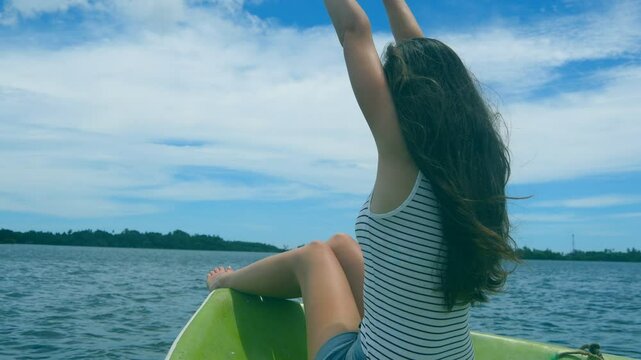 Happy woman sits on bow of boat and admires a beautiful nature landscape during trip. Pretty girl with blowing long hair rests on deck of yacht and enjoys summer travel. Vacation or holiday concept