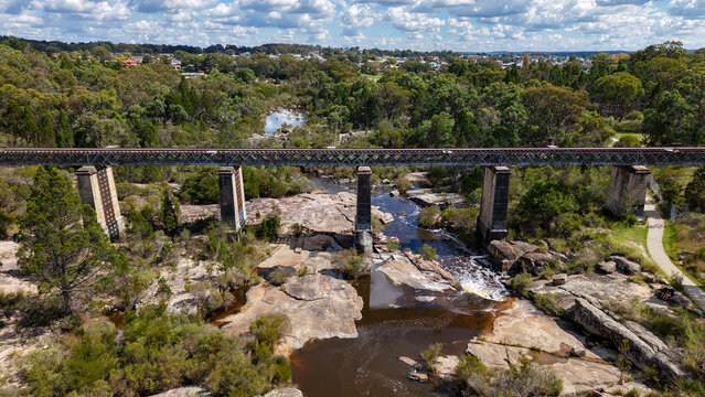 Old red railway bridge in stanthorpe stretching over rocky riverbed through forest landscape under cloudy sky