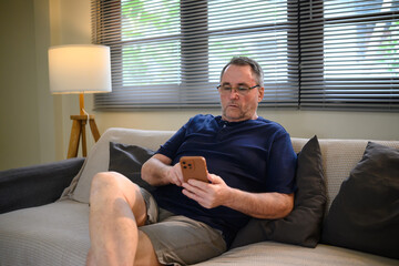 Obraz premium Senior man relaxing on sofa and using smartphone at home
