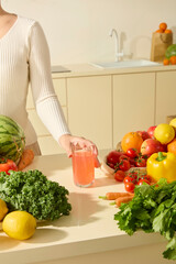 A woman is placing a glass of watermelon juice on a kitchen island filled with colorful vegetables: lemons, apples, tomatoes, celery, cauliflower... Minimalist, modern kitchen space
