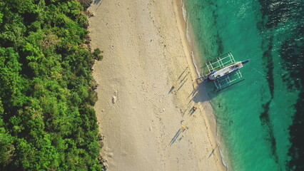Aerial drone view of a tropical beach with turquoise water and white sand meeting a lush green forest, with a traditional outrigger boat anchored near the shore