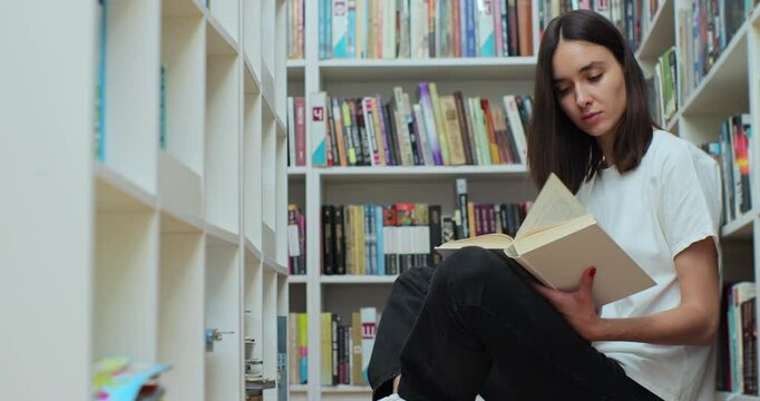 Beautiful girl student reading book, sitting among shelves with book on library floor, side view. - Powered by Adobe