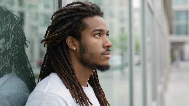 Young man with dreadlocks thinking by glass wall outside	