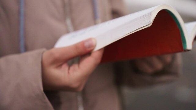 Close-up view of lady's hands gently flipping pages of red book in autumn setting, blurred background with soft coat fabric visible, capturing peaceful reading moment in natural outdoor light