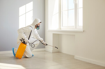 Professional exterminator in protective white suit and gas mask spraying insecticide on floor in empty room. Male pest control worker with sprayer bottle using chemical treatment for disinfection.