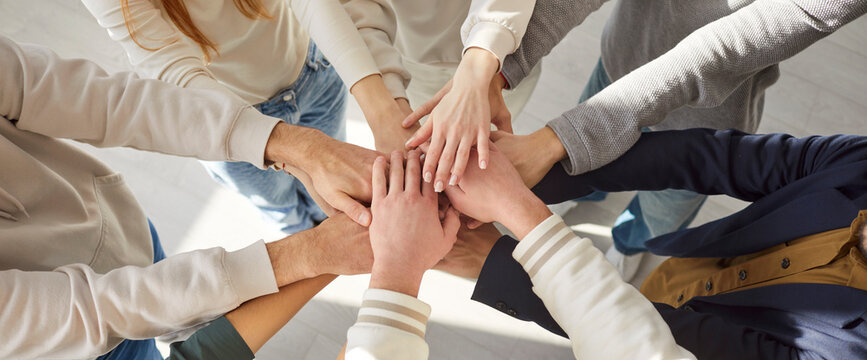 Closeup top view of people group in casual clothes putting their arms in a pile standing indoors. Students gathered together making hands in stack. Friendship, unity and teamwork concept. Banner.