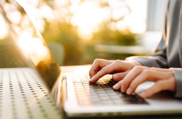 Hands of young woman typing on laptop keyboard in sunset rays. Close-up of hands of female...