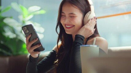 Happy asian woman using smartphone with wireless headphones at modern coffee shop, reusable cup on table – digital lifestyle, eco-conscious living and urban comfort