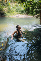 Fototapeta premium woman enjoys a serene moment while sitting in a clear, natural pool, surrounded by vibrant green foliage. peaceful water flows gently over smooth rocks under the warm sun