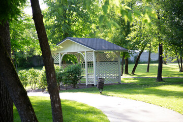a white wooden gazebo with lattice walls and a dark roof, located in the middle of a green park. A...