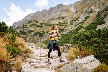 Young female hiker with yellow hiking backpack on rocky trail with mountain view. Happy woman enjoying weather and outdoor adventure. Travel concept.