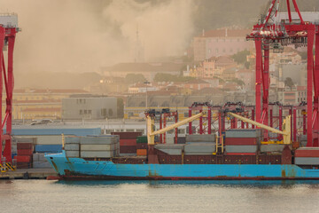 Container ship and red cranes at Lisbon port with cityscape and fire