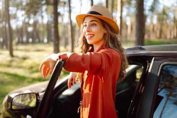 Happy young woman stands near open car door enjoying sunset in forest. Back view of female traveler...